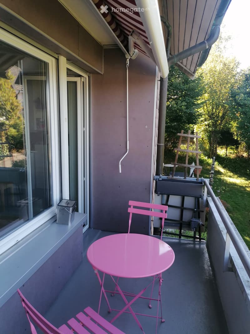 Balcony with pink metal table and chairs, overlooking a green area with trees.