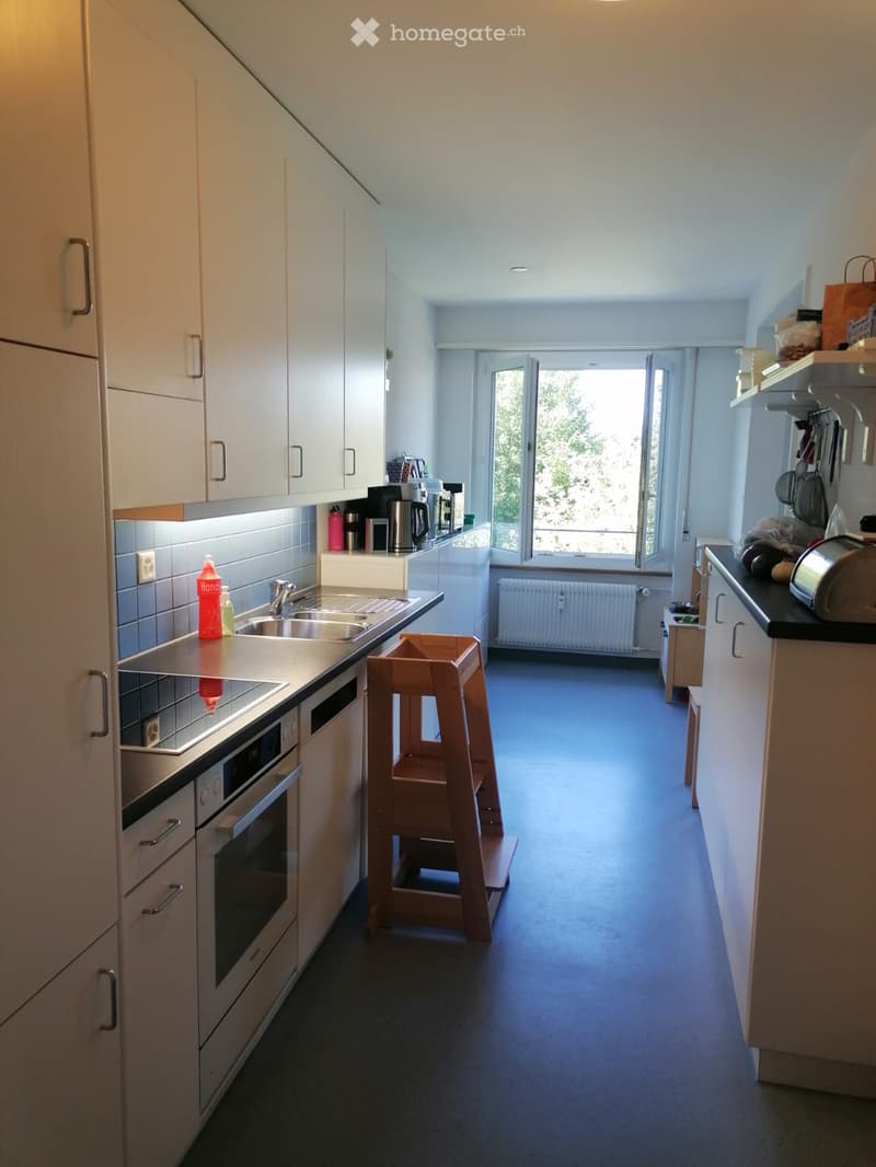 Modern kitchen with white cabinets, stainless steel appliances, a sink, and a window overlooking greenery. Includes a wooden stool, coffee maker, and various kitchen items on the counter.