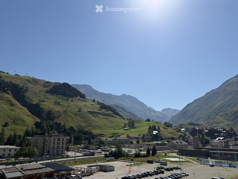 A scenic mountain valley with a town, parking lot, and surrounding green hills and peaks.