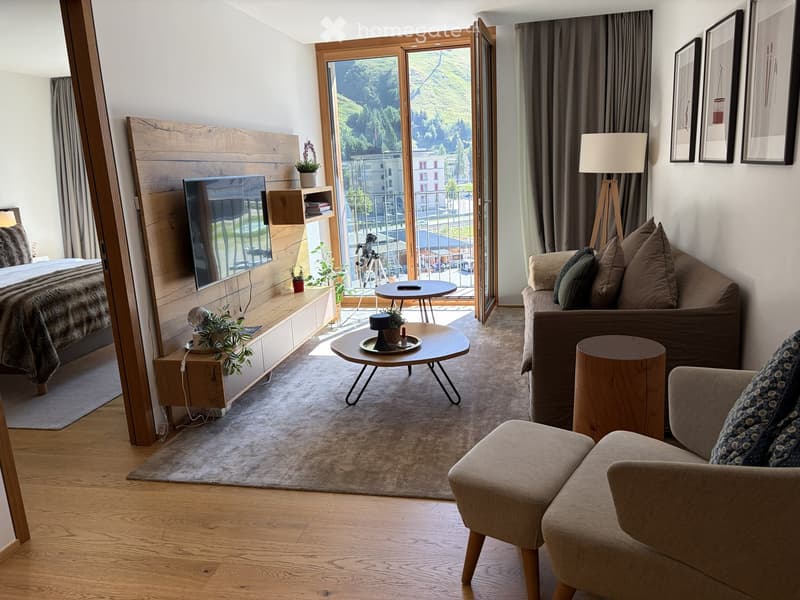 Modern furnished room with a sofa, coffee table, armchair, bed, small table, lamp, and framed picture on the wall. Large windows offer a scenic view.