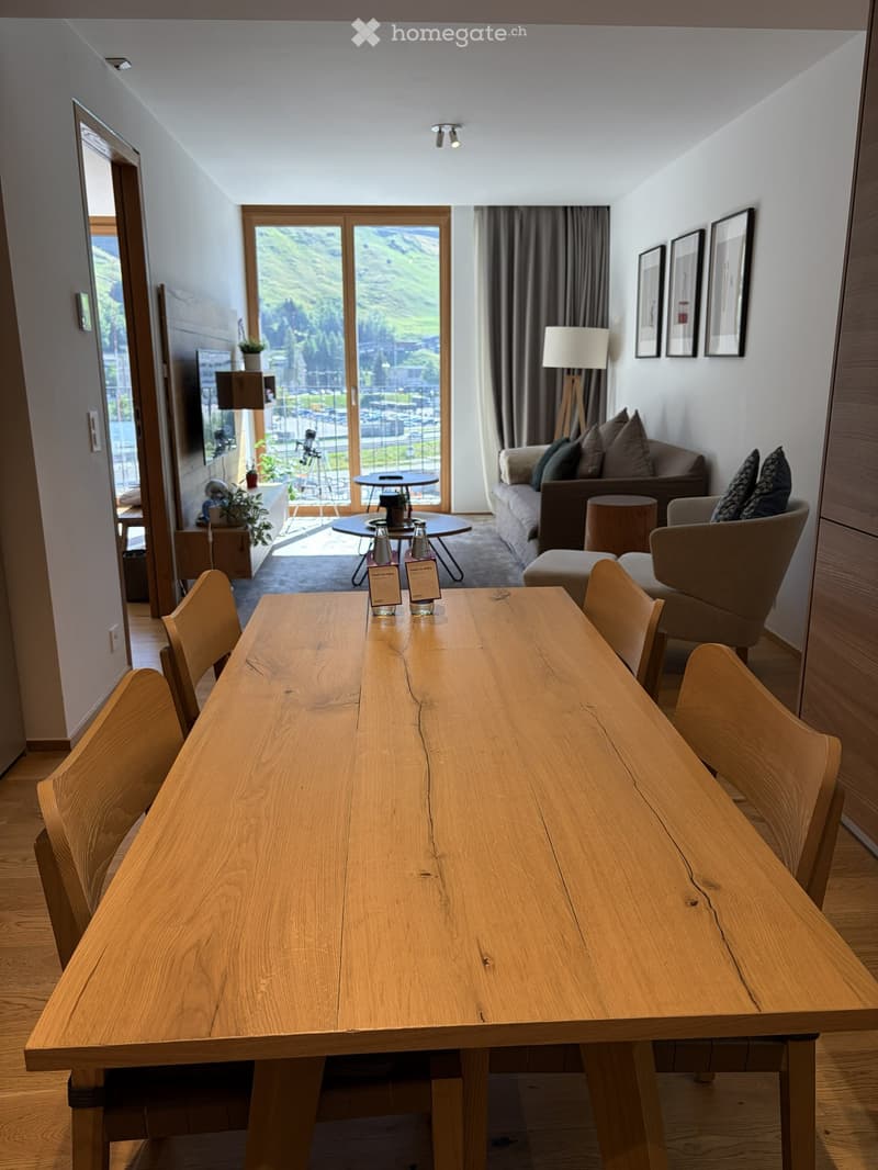 Modern living and dining area with a large wooden dining table, chairs, sofa, and a view of the mountains through large windows.