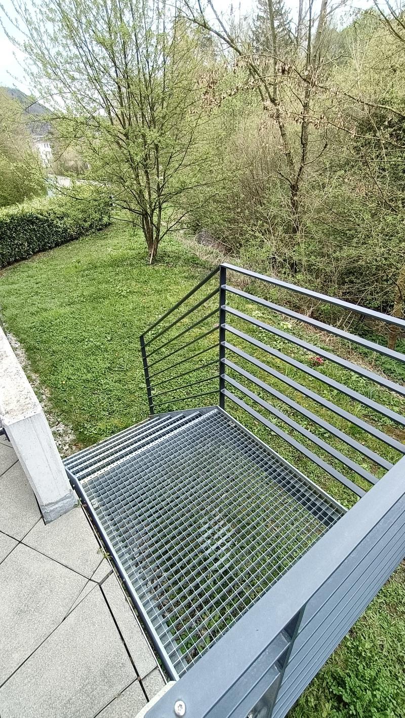 Metal staircase with a grid-like railing leading down to a grassy area surrounded by trees.