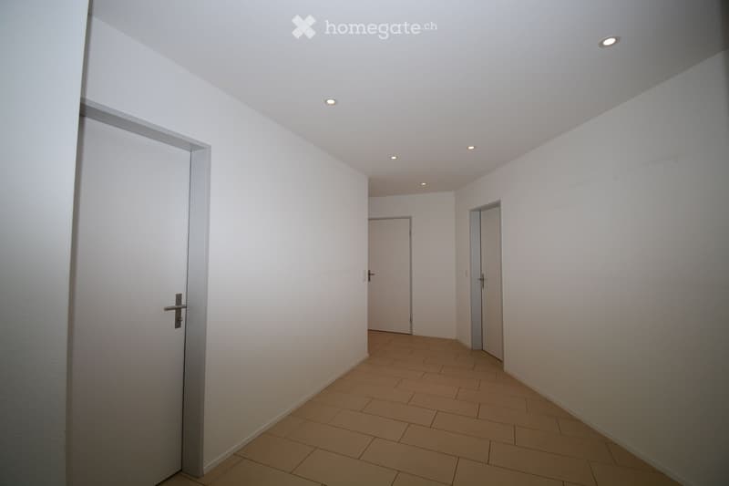 Empty hallway with white walls, beige tiled floor, and recessed ceiling lights. Three doors are visible, two on the right and one on the left.