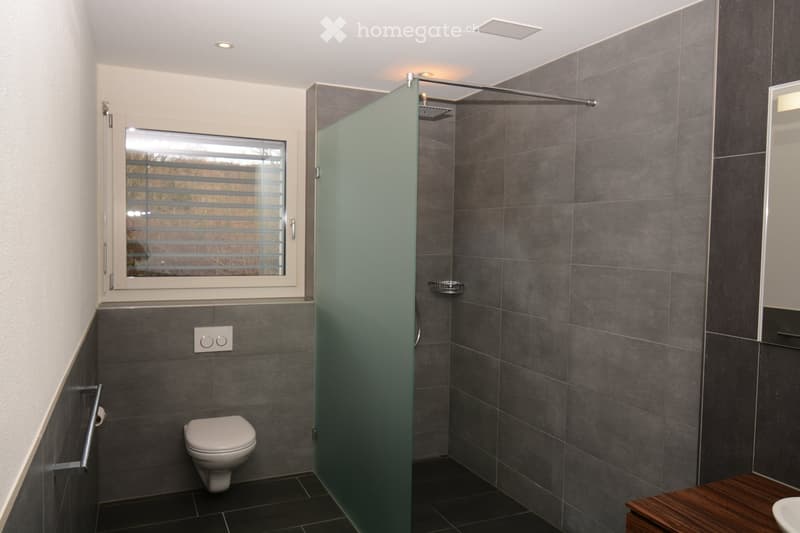 Modern bathroom with dark grey tiles, a white toilet, a glass-enclosed shower, a wooden bench, and a window with blinds.
