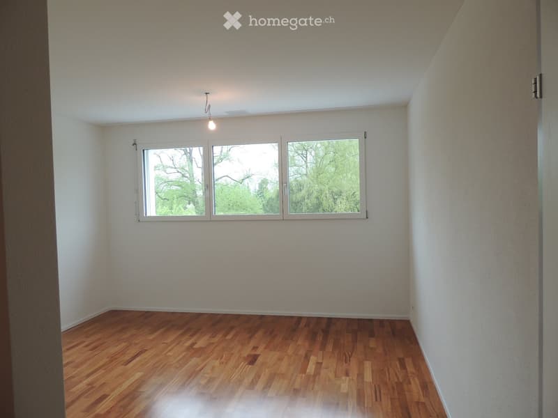 Empty room with wooden flooring, white walls, and large windows showing greenery outside.