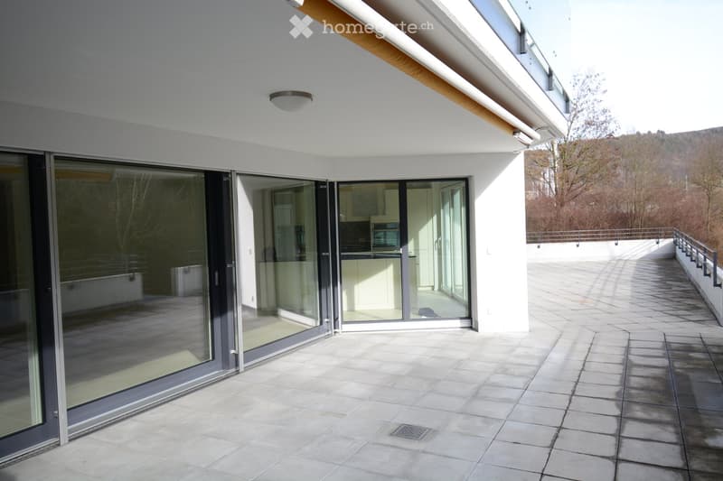 Modern balcony with large glass sliding doors leading to a kitchen area, tiled floor, and a scenic view of a wooded area.