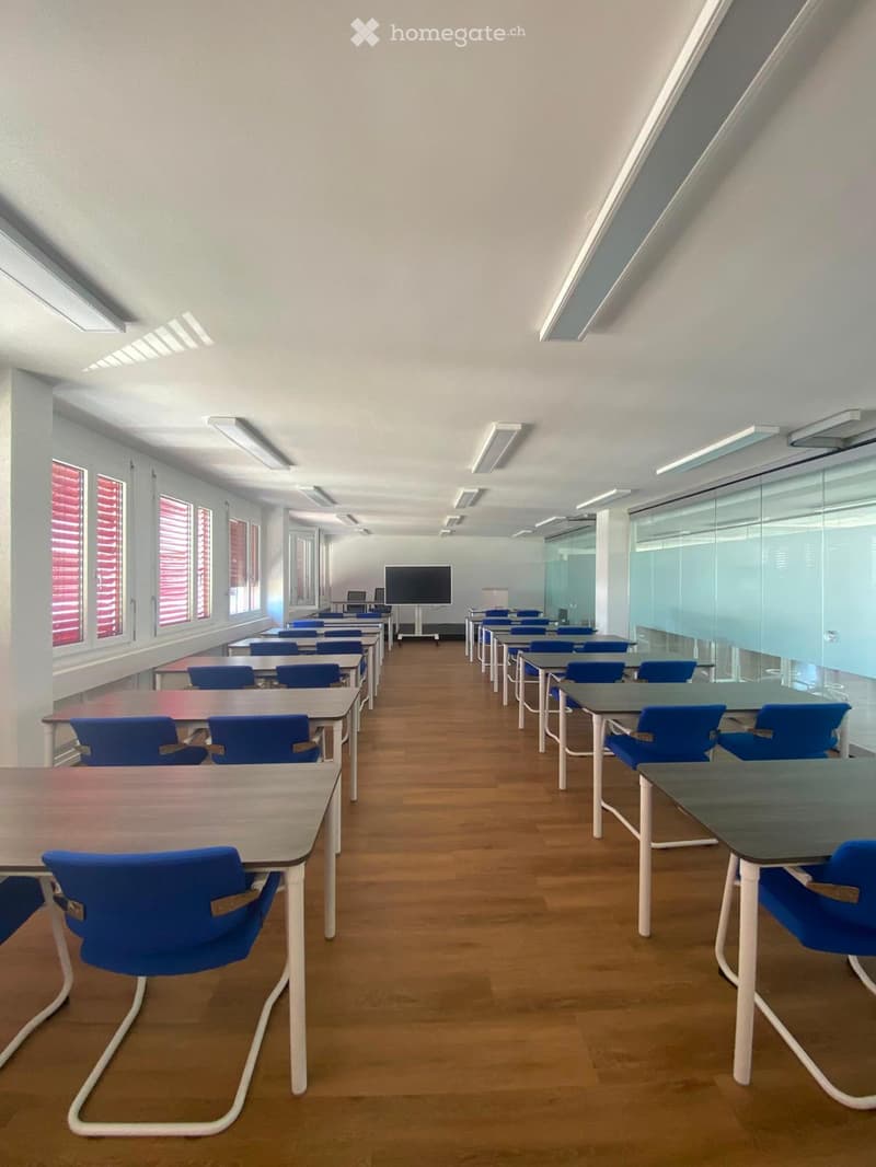 Classroom with wooden flooring, multiple tables and chairs arranged in rows, large windows with red blinds, and a screen at the front.