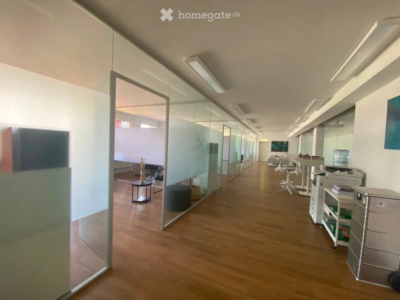Modern hallway with wooden flooring, glass partitions, and visible office furniture in the background.