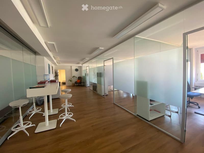Modern office space with wooden flooring, white stools, glass partitions, and a small seating area with a chair and table.