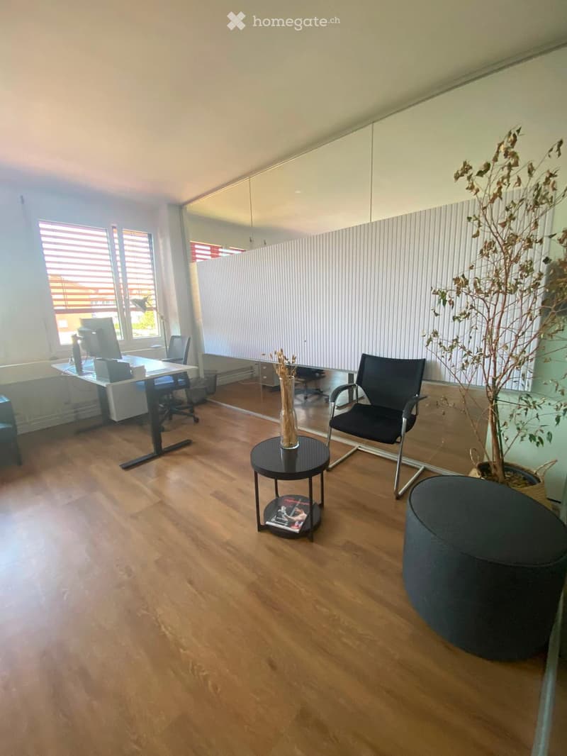 Modern office space with wooden flooring, large windows, a desk with a computer, a round table with a vase, and a comfortable chair.