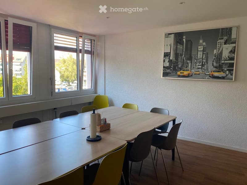 Bright room with large windows, a long rectangular table surrounded by chairs, and a large framed cityscape picture on the wall.