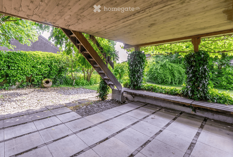 concrete patio with a wooden deck, a staircase leading up, a concrete bench, greenery around