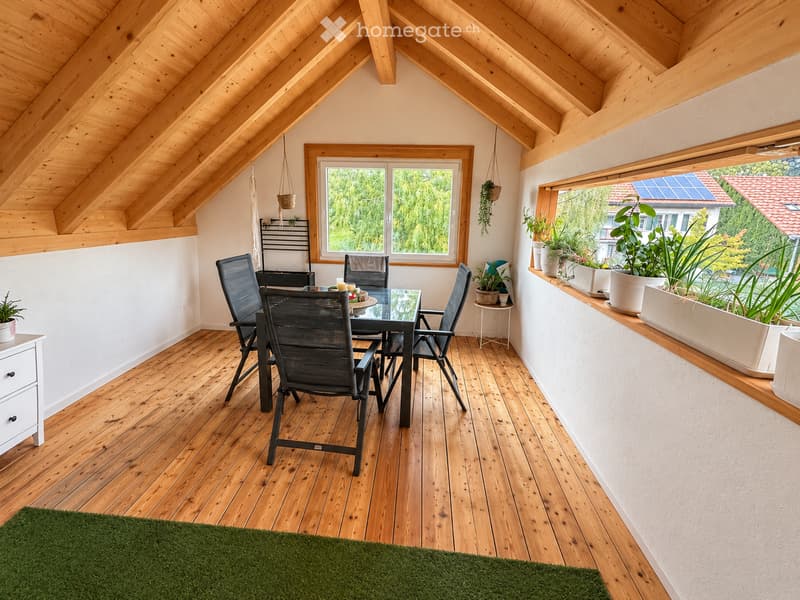 Bright, wooden-floored room with slanted wooden ceiling, large windows, dining table with chairs, side table, potted plants, and a green artificial grass rug.