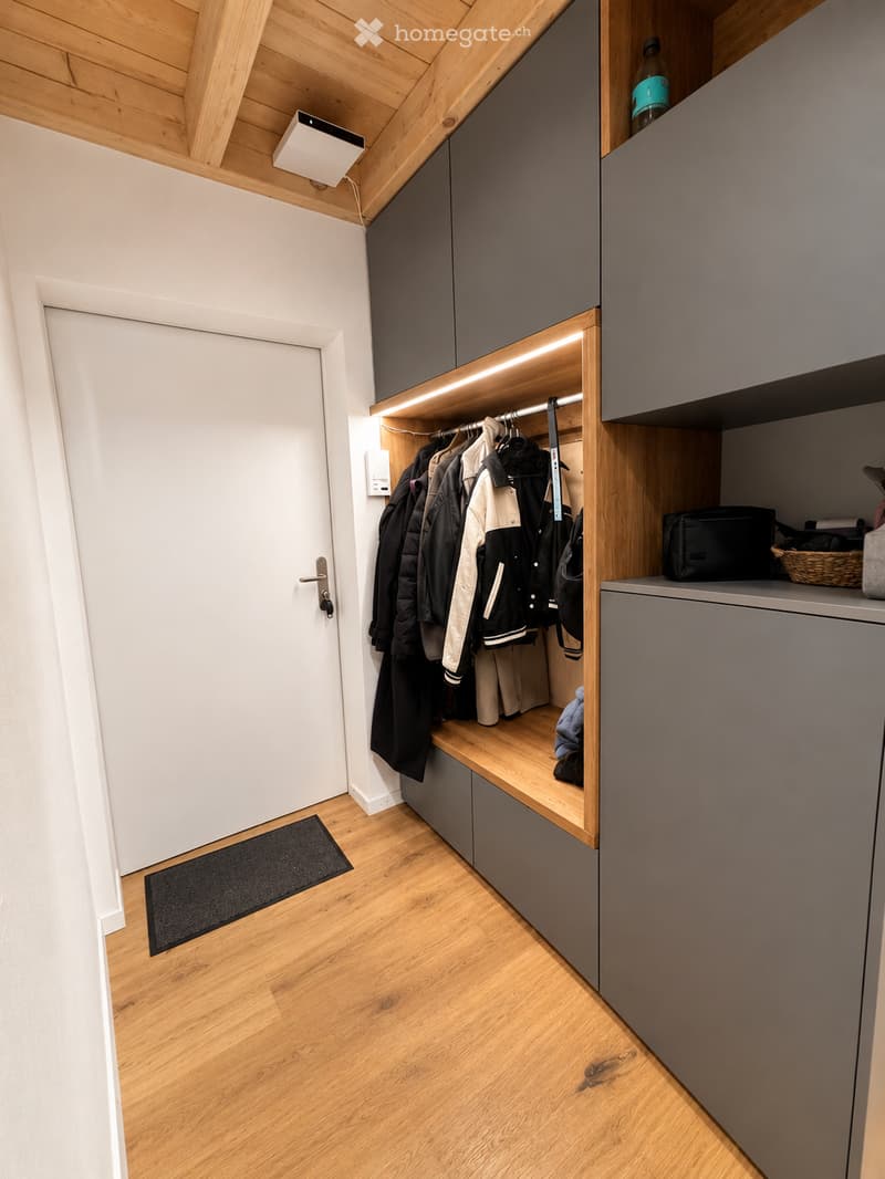 Modern hallway with wooden flooring, built-in storage unit with coats and bags, grey cabinets, and a small bench.