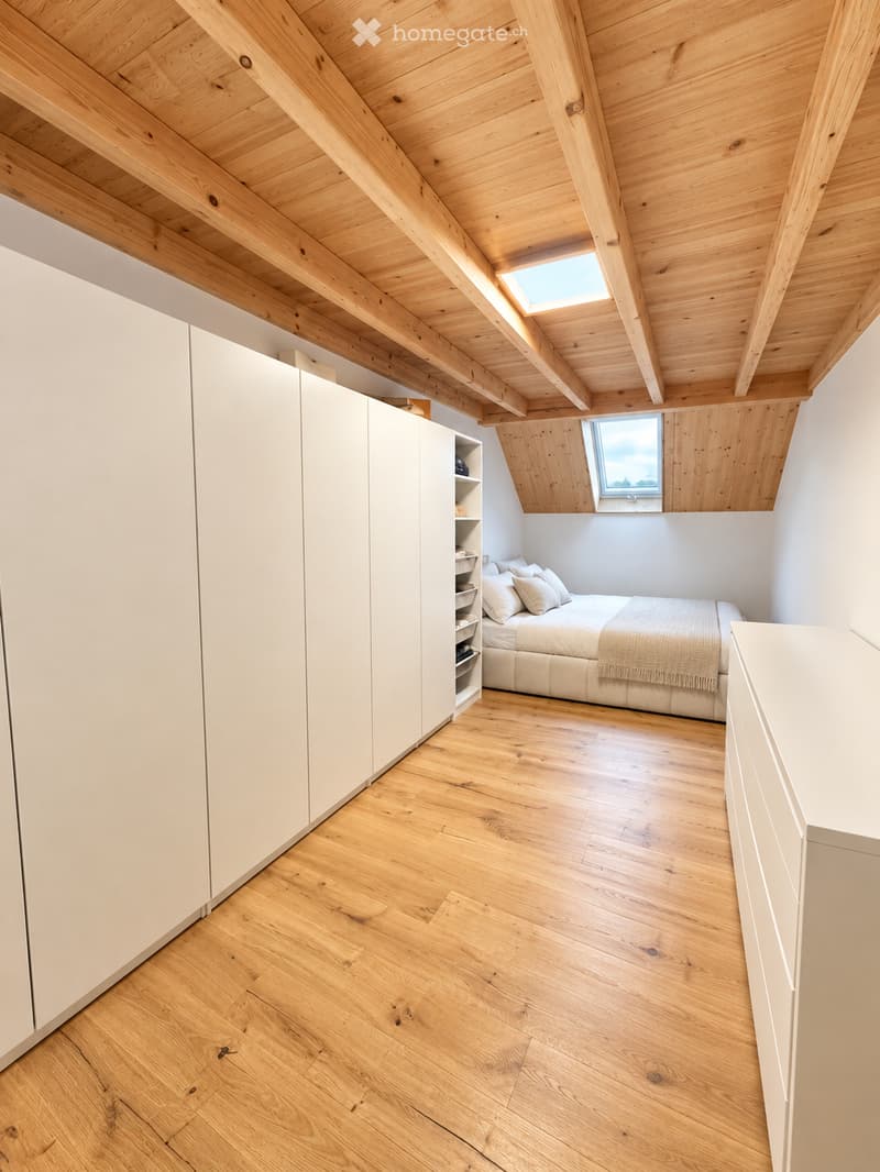 Bright, modern bedroom with wooden floors, slanted wooden ceiling, skylight windows, and built-in white wardrobe and shelving unit.
