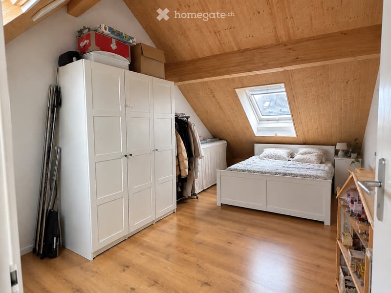Bright attic bedroom with wooden floors, slanted wooden ceiling, skylight window, bed with pillows and duvet, large white wardrobe, nightstand with lamp, toy shelf, and skis leaning against the wall.