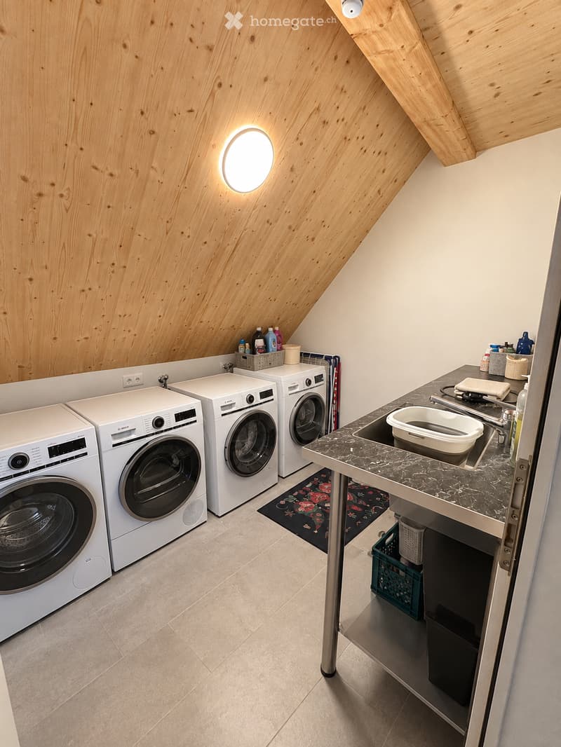 Modern bathroom with wooden slanted ceiling, white tiled floor, and marble countertop. Features a sink, toilet, washing machine, and dishwasher.