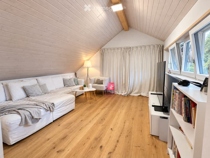 Bright living room with wooden floors, white sofa, coffee table, armchair, bookshelf, and TV. Features large windows and a slanted wooden ceiling.