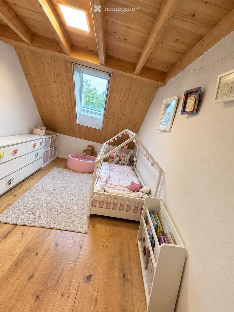 Cozy children's bedroom with wooden slanted ceilings, large window, bed with pink bedding, bookshelf, drawer unit, and a small rug.