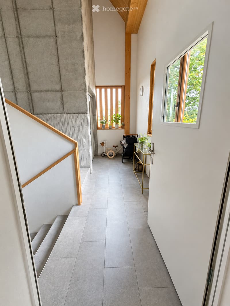 Modern hallway with grey tiled floor, white walls, wooden staircase railing, and a small window showing greenery outside. A small table with plants and a stroller is visible near the window.