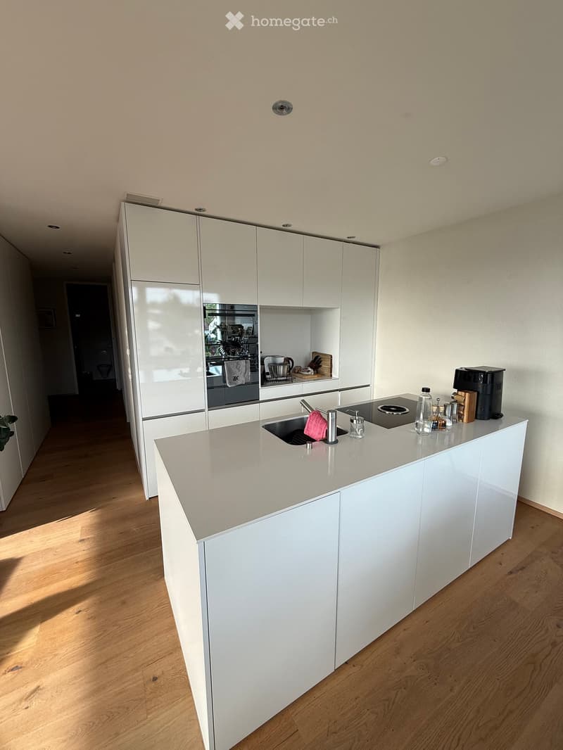 Modern kitchen with white cabinets, light grey countertop, sink, coffee maker, and built-in oven. Wooden flooring and bright lighting.