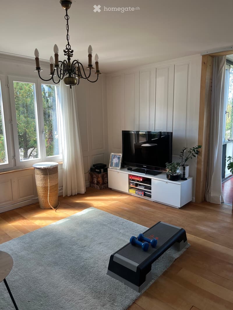 Bright living room with wooden flooring, large windows, white cabinetry, television, exercise bench with dumbbells, and a chandelier.