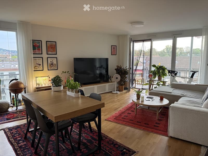 Bright living room with large windows and a view, featuring a grey sectional sofa, coffee table, dining table with chairs, TV, fan, globe, and potted plants.