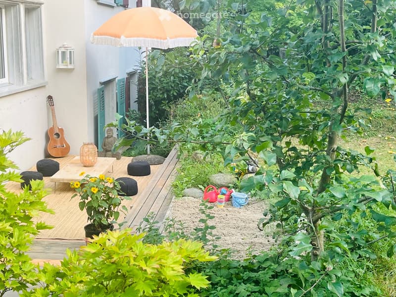 A serene garden area with a wooden deck, small table, cushions, a guitar, a lantern, a small statue, and a play area with toys and sand.