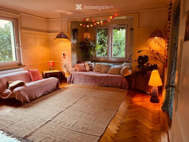 Cozy living space with a daybed, sofa, small table, and warm lighting. Features wooden flooring, large windows with greenery outside, and decorative plants.
