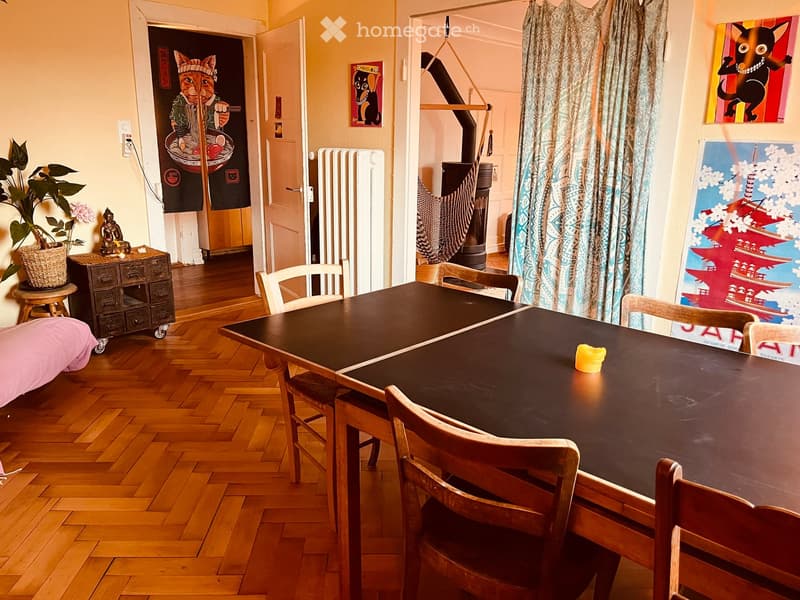 Dining area with a large wooden table surrounded by chairs, a small cabinet with drawers, a potted plant, and colorful artwork on the walls.