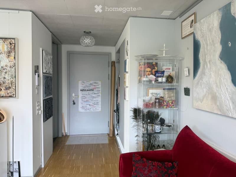 A modern hallway with a red sofa, a glass cabinet, and artwork on the walls. The space features wooden flooring and a chandelier.