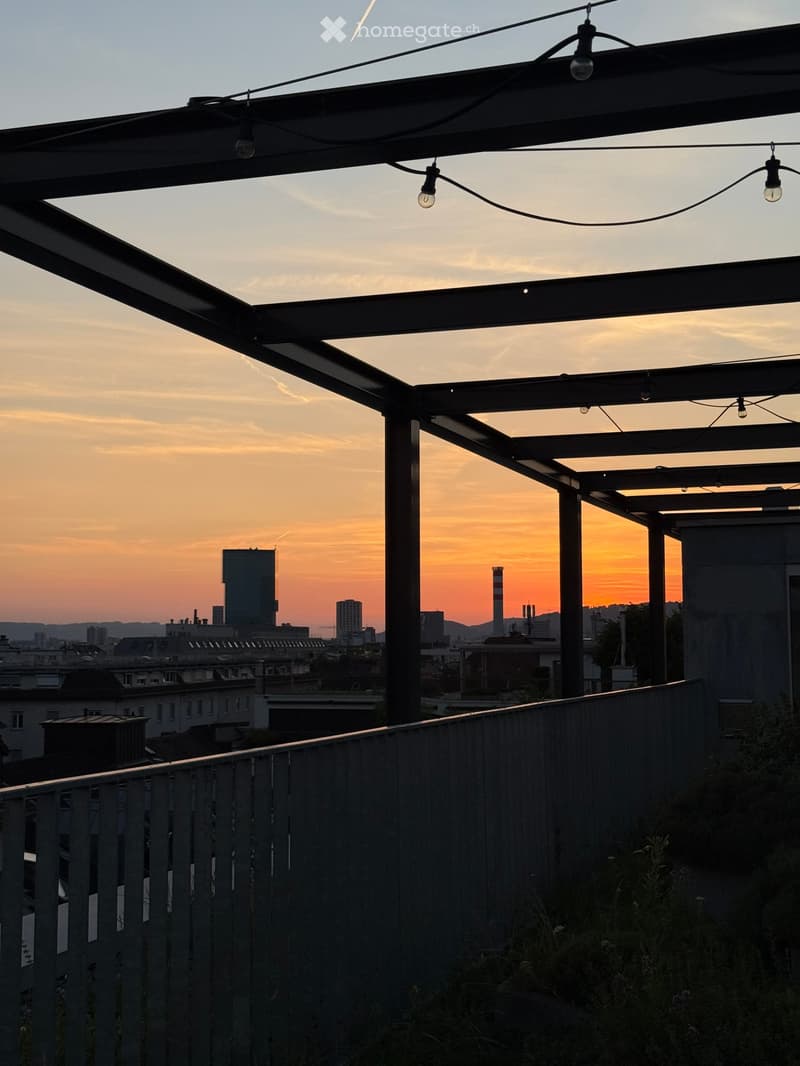 Roof terrace with metal railing, string lights, and a cityscape view at sunset.