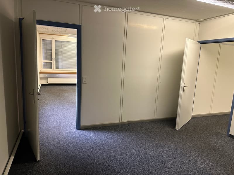 Empty room with grey carpeted floor, white walls, and open doors leading to adjacent spaces. No furniture or equipment visible.