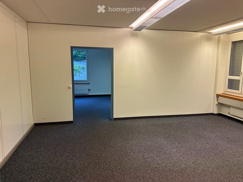 Empty room with white walls, carpeted floor, and a door leading to another space. There is a small window with blinds on the right side.