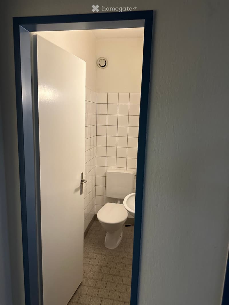 Small bathroom with white tiled walls, a toilet, and a sink, viewed through an open door.