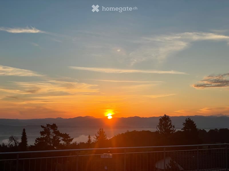 Balcony overlooking a scenic sunset view of mountains and a lake, with trees in the foreground and a metal railing.