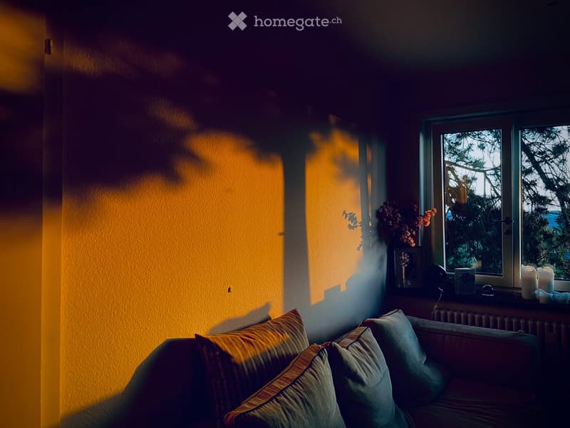 A cozy living room with a sofa, pillows, a small table with a vase of flowers, a picture frame, and candles on the windowsill. The room has warm yellow walls and a window showing trees outside.