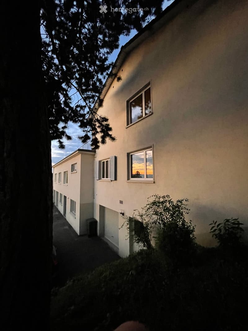 Two-story residential building with white walls, multiple windows, and a small balcony visible on the upper level.