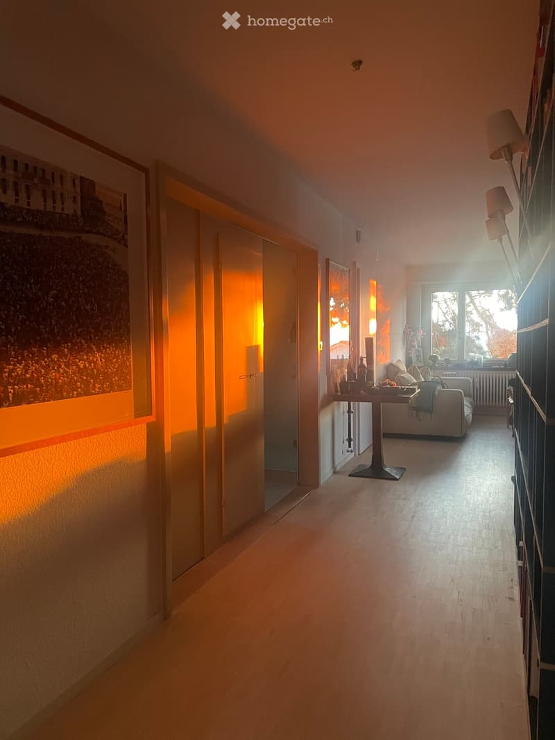 A well-lit hallway with wooden flooring, leading to a living room area featuring a sofa, coffee table, and a bookshelf. The hallway has a large framed picture on the left wall and a glass door at the end.