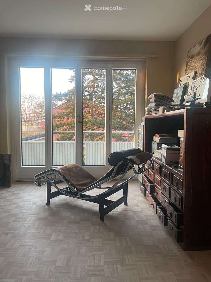 A modern room with wooden flooring, large windows, and a sleek black lounge chair. A wooden cabinet with drawers and shelves holds books and decorative items.