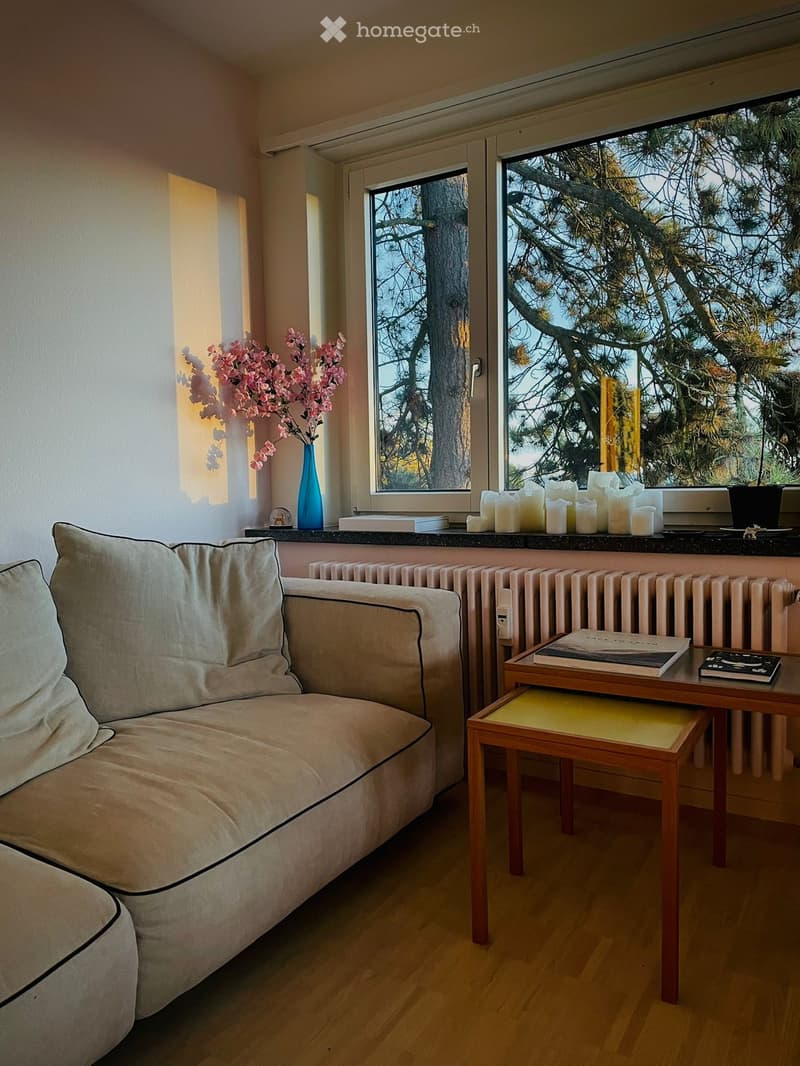 A cozy living room with a light-colored sofa, a small wooden table with a stool, a vase with pink flowers, and several candles on the windowsill. The room has wooden flooring and large windows offering a view of trees.