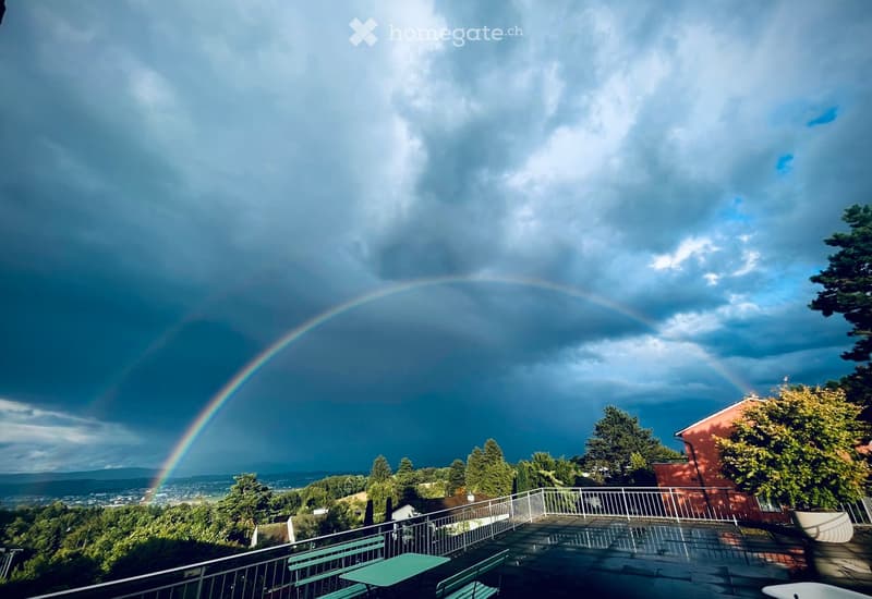 A balcony or terrace with a scenic view of a city and surrounding hills under a partly cloudy sky, featuring a rainbow. The balcony has a metal railing, a green table, and green chairs.