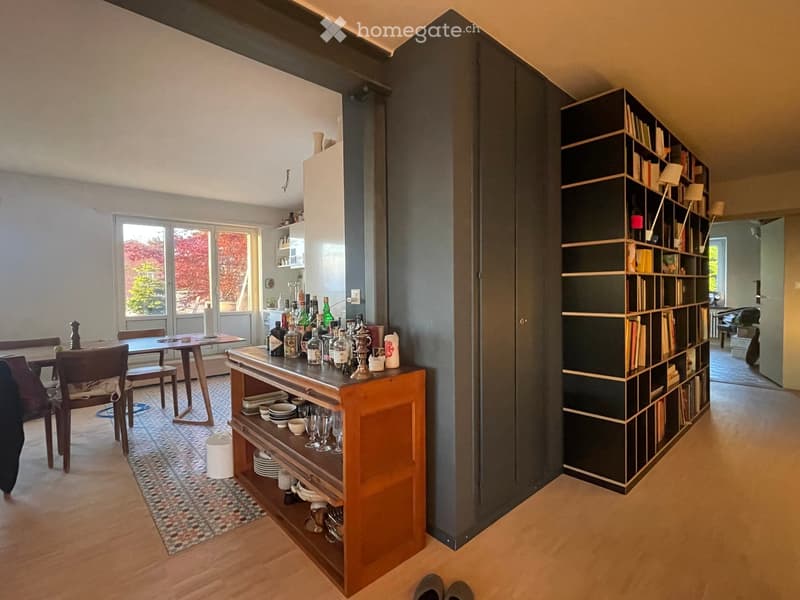 A modern, open-concept living and dining area with wooden flooring, a large bookshelf, a dining table with chairs, and a wooden cabinet holding wine bottles, plates, and glasses.