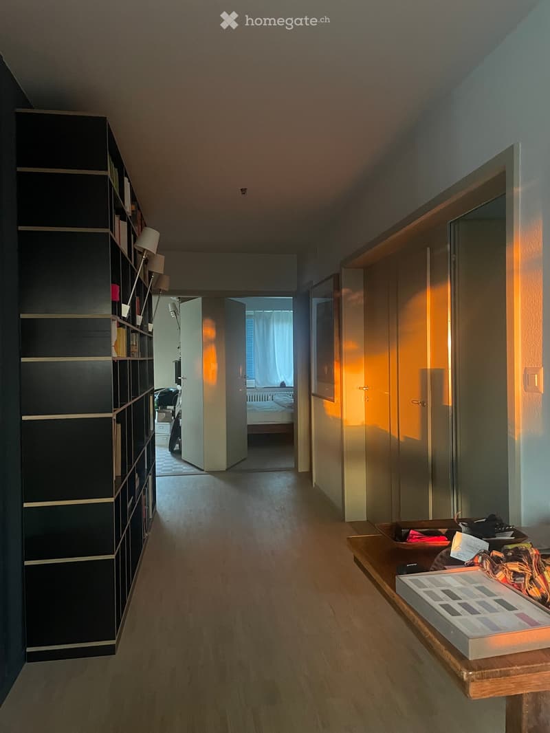 Modern hallway with wooden flooring, built-in shelving unit on the left, and a small table with items on the right. Open doors lead to other rooms, including a bedroom visible in the background.