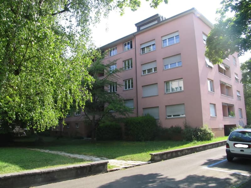 A multi-story residential building with pink facade and multiple windows, surrounded by greenery and a paved pathway.