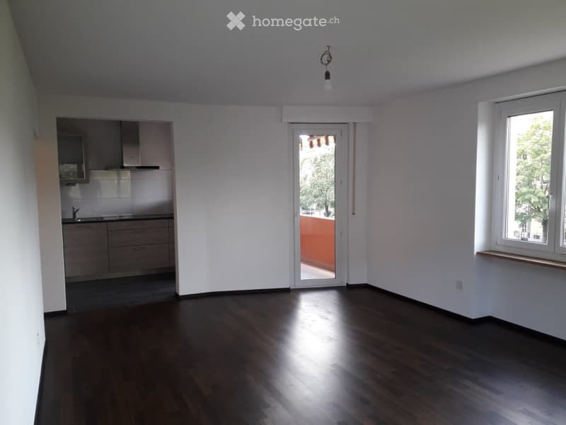 Empty room with dark wooden flooring, white walls, and a sliding glass door leading to a balcony. A small kitchen area is visible in the background.