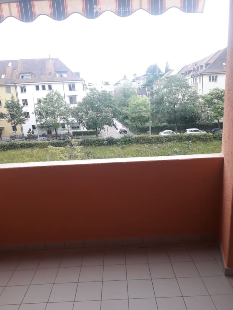 Balcony with tiled floor, striped awning, overlooking a street with houses, trees, and parked cars.