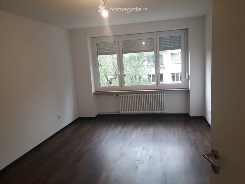 Empty room with wooden flooring, large window with blinds, radiator, and a hanging light fixture.