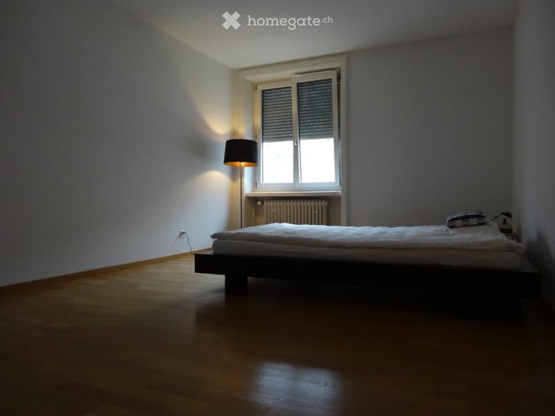 Minimalist bedroom with wooden flooring, a single bed, a nightstand, and a floor lamp near a window with blinds.