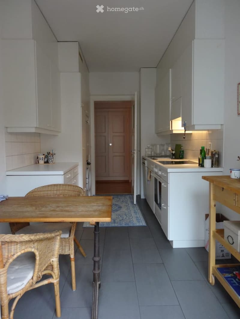 Modern kitchen with white cabinets, a wooden dining table, wicker chairs, and a small open area leading to another room.